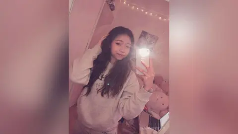 Young woman in a cozy grey sweatshirt taking a mirror selfie in a warm, pink-lit bedroom setting.