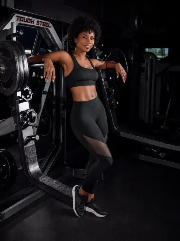 KylieDecks — KylieDecks poses in a black sports bra and leggings while leaning against a weight machine in a gym.