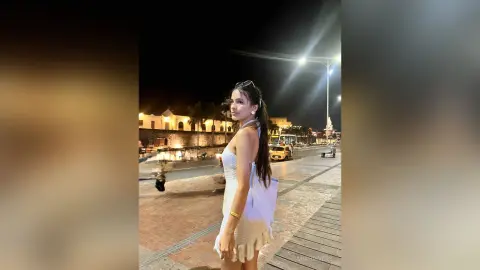 Young woman in a white summer dress walking on a boardwalk at night near city lights.