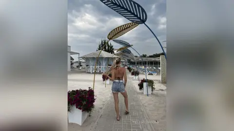 Blonde woman in denim shorts and bikini top walking through a tropical beach resort walkway under decorative leaf arches.