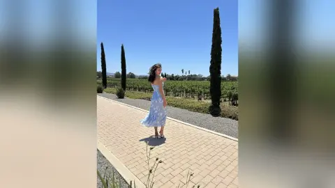 Young woman in a blue floral sundress walking through a sunny vineyard path lined with tall cypress trees.