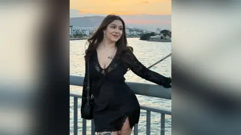 Smiling woman in a black lace dress enjoying a sunset view by the waterfront.