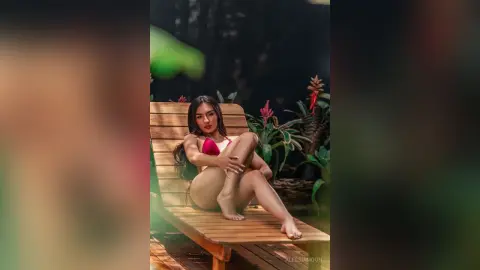 Glamorous woman in a red bikini relaxing on a wooden lounge chair in a lush tropical garden setting.