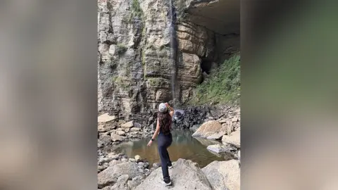 RossiNoleen stands on a rocky ledge in front of a tall waterfall, wearing a black tank top and black leggings.