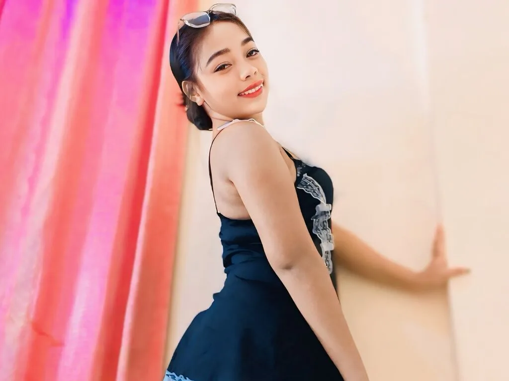 MissyKim smiles while wearing a black tank top and glasses, posing against a light wall next to pink curtains.