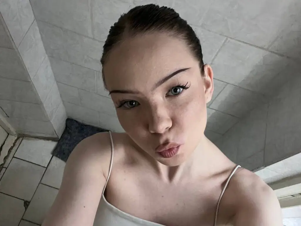 Close-up selfie of a young woman with natural makeup in a white camisole against a tiled bathroom background.