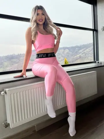 Smiling woman in pink gym set sitting on a windowsill overlooking a scenic mountain landscape.