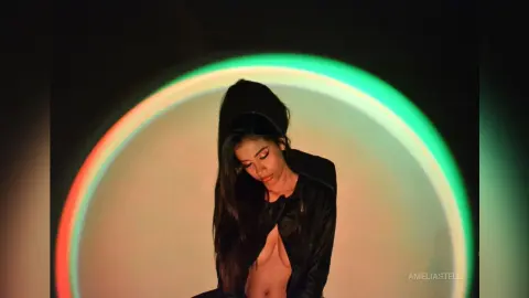 AmeliaStell looks down with long dark hair while wearing a black open-front top against a bright circular light background.