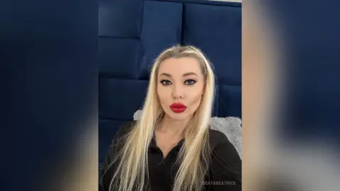 Blonde woman with red lipstick in a black outfit posing against a navy blue velvet headboard.