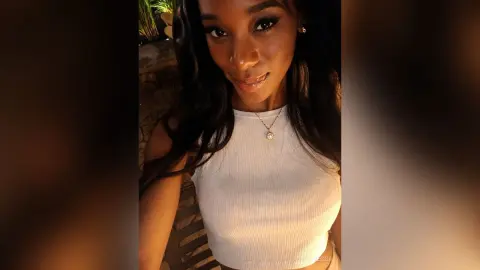 Smiling Black woman in a white ribbed halter top wearing a gold necklace in a warm evening outdoor setting.
