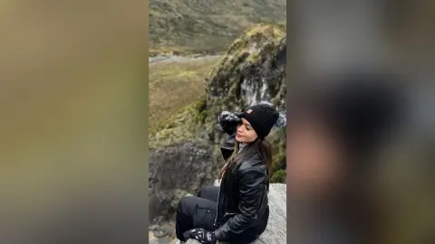 Woman in black leather jacket and beanie sitting on a mountain cliff overlooking a scenic valley landscape.