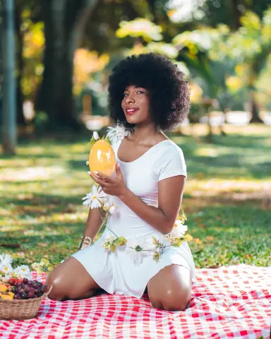 Smiling woman in a white floral dress holding a yellow egg during a sunny garden picnic on a red checkered blanket.
