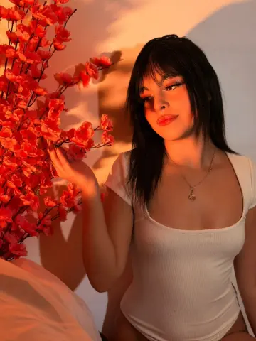 DapneOwen wears a white bodysuit and a necklace while posing next to bright pink flowers.