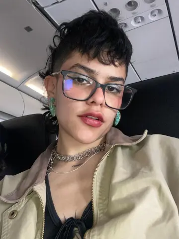 Young woman with short dark hair and glasses wearing a beige jacket and silver necklaces on an airplane.