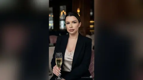 CapriAndersen poses in a black blazer and white lace top, holding a glass of champagne with her dark hair slicked back.