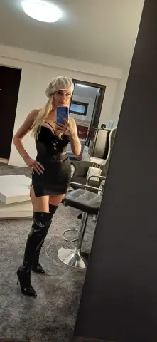 Blonde woman in a black bodysuit and tall patent leather boots taking a mirror selfie in a modern room.