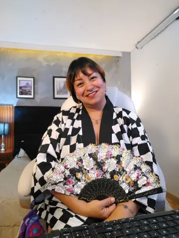 Smiling woman in a black and white checkered kimono holding a decorative floral hand fan in a bedroom setting.