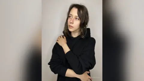 Young woman in a black hoodie posing thoughtfully against a neutral studio background.