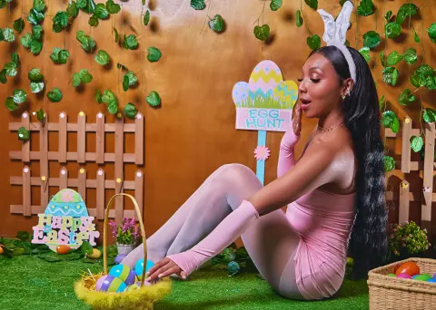 Woman in pink outfit sitting by Easter decorations and egg hunt sign in a festive spring setting.