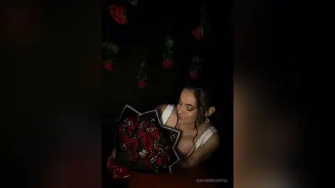 NahomiRomero holds a large bouquet of red roses against her chest, wearing a white top with her dark hair pulled back in a dark setting.