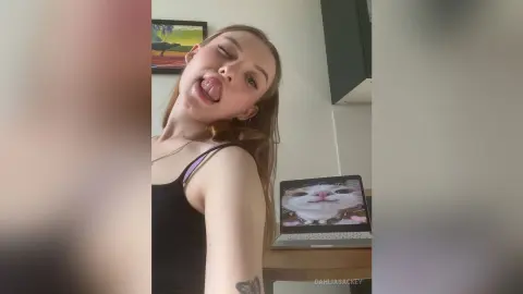 DahliaSackey sticks her tongue out while wearing a black tank top, sitting near a laptop in a kitchen setting.