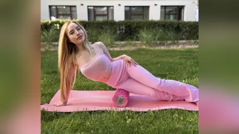 Blonde woman in pink ribbed yoga outfit relaxing on a mat in a sunny green grass field.
