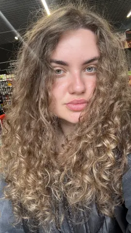 Young woman with long curly blonde hair wearing a black leather jacket in a grocery store setting.