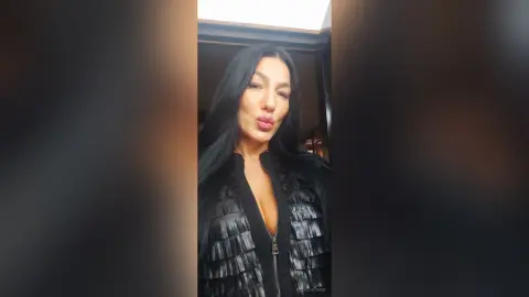 Glamorous woman in a black textured jacket posing with a confident expression in a dimly lit indoor setting.