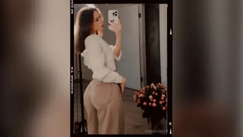 TarenLeonetti poses in a white long-sleeve sweater and tan pants, showing her curves in a mirror selfie with long brown hair.