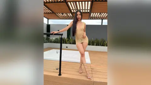 IrinaRussel stands on a wooden deck wearing a tan, open-knit crochet dress and strappy sandals, posing next to a white hot tub under a wooden pergola.
