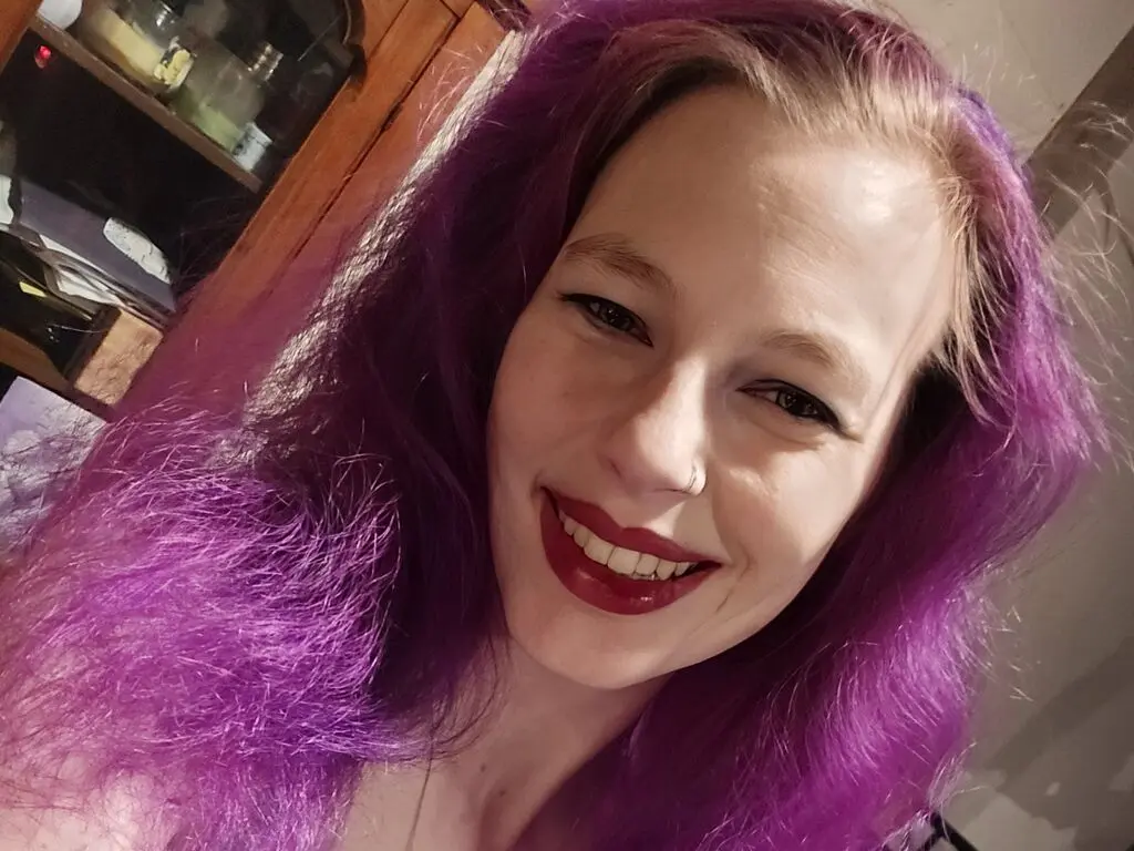 Smiling woman with vibrant purple hair in a cozy indoor setting.