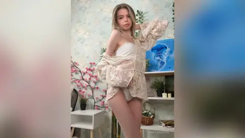 UnataMigdel poses in a white lace outfit and panties, looking at the camera in her bedroom.