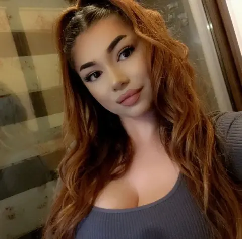 Young woman with long wavy auburn hair wearing a grey ribbed top in a cozy indoor setting.