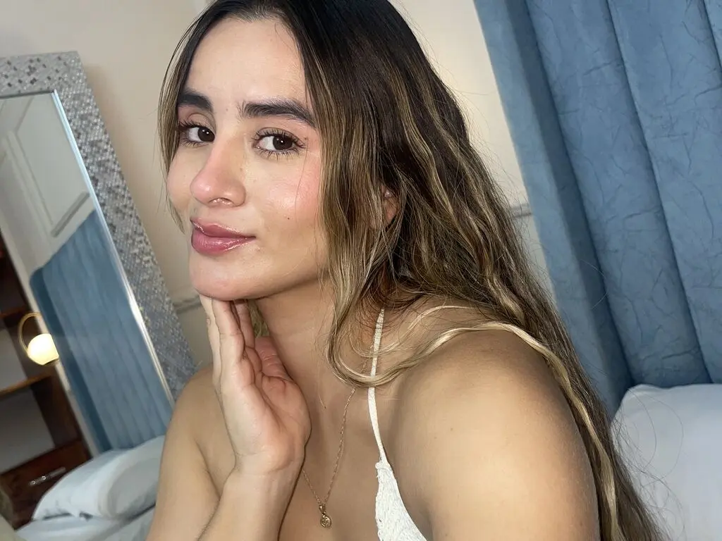 AliceChanell poses with her hand under her chin, wearing a white lace top and a gold necklace, with her long wavy hair falling over her shoulder.