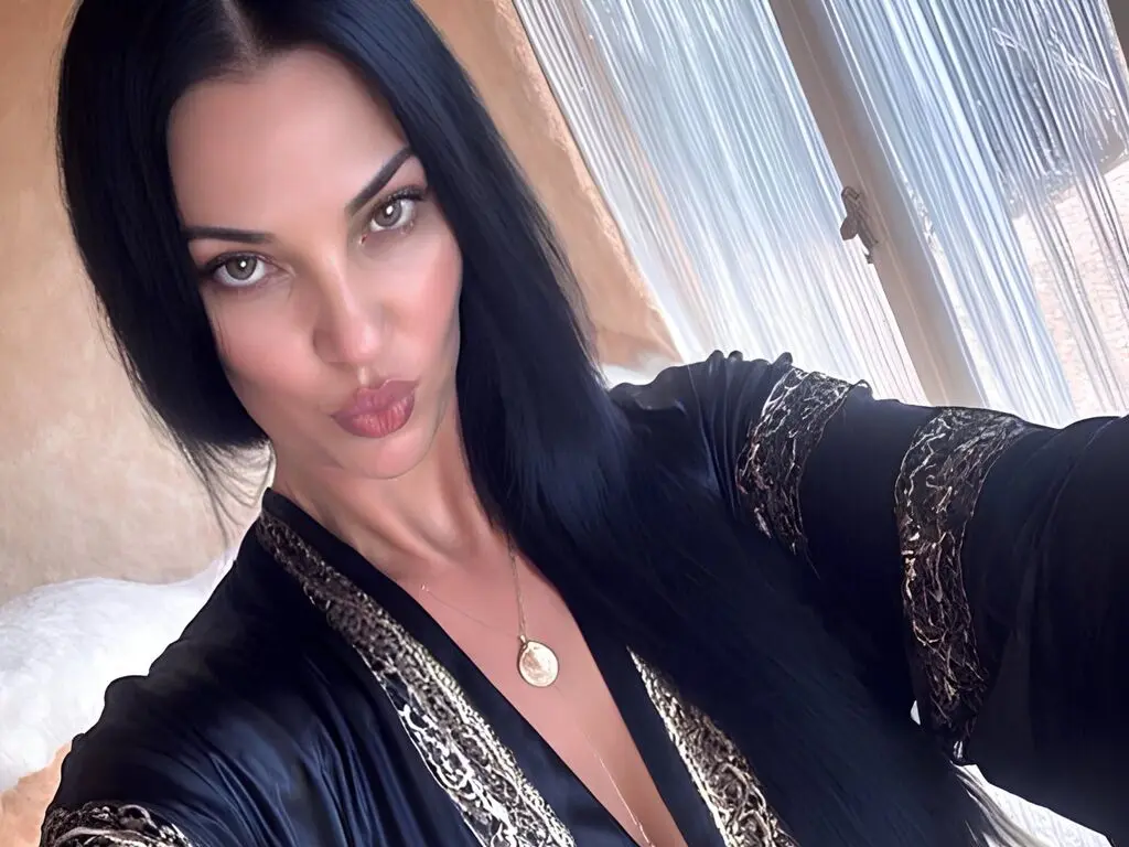 Woman with long black hair wearing a black and gold patterned robe in a bright, sunlit room.
