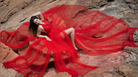 NataliaNacher poses on red rocks wearing a long, flowing red dress that spreads out around her body.