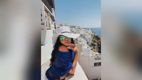 MissyJolie wears a white sun hat and blue mesh top while posing on a white balcony overlooking a coastal town.