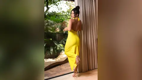 Elegant woman in a bright yellow backless evening gown posing in a lush tropical garden setting.