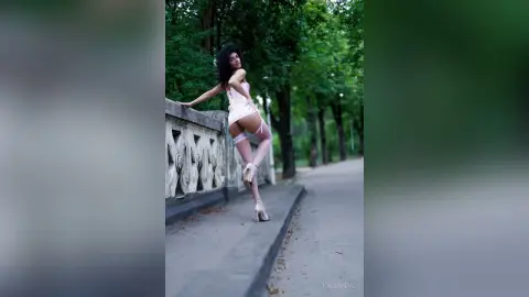 Curvy woman in a pink satin slip dress and white stockings posing on a stone balcony in a lush green park.