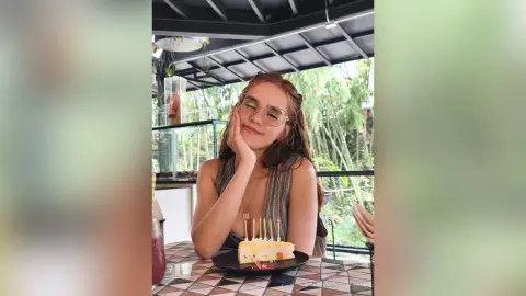 AmelieLautrec wearing sunglasses and a brown sleeveless top, resting her chin on her hand while sitting at a table with a small cake.