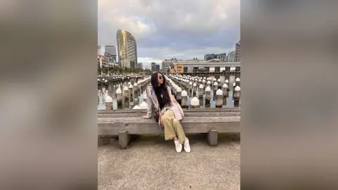 Stylish woman in a beige trench coat sitting on a wooden pier by a city waterfront under a cloudy sky.