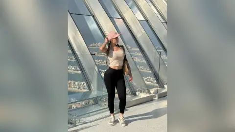 Stylish woman in black leggings and beige crop top posing by a glass window overlooking a coastal city view.