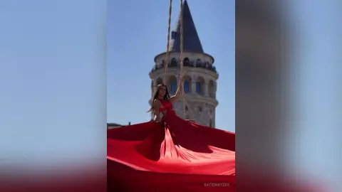 Elegant woman in a flowing red gown sitting on a swing in front of the historic Galata Tower under a blue sky.