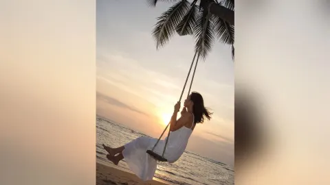 MilaSwan — MilaSwan sits on a swing suspended from a palm tree over the beach at sunset wearing a white dress.