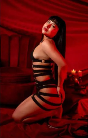 Glamorous woman in black strappy lingerie posing in a moody red velvet bedroom setting.