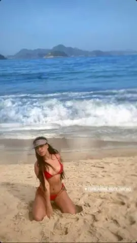 Young woman in a red bikini relaxing on a sunny beach with ocean waves in the background.