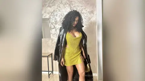 NicoleBella wears a tight, short yellow glitter dress and a long black shiny coat while standing in a room.