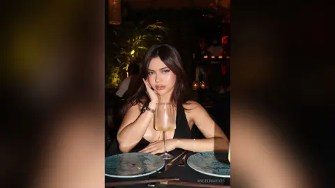 Young woman in a black dress sitting at a dimly lit restaurant table with a glass of white wine.