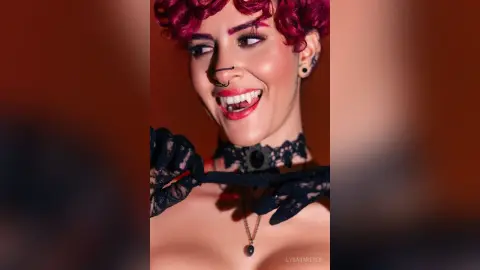 LysaraReyes smiles with red hair and red lipstick, wearing a black lace choker and black lace gloves against a warm orange background.