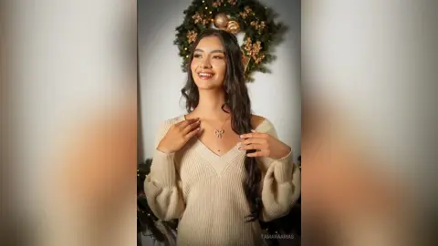 Smiling young woman in a cream ribbed sweater posing in front of a festive Christmas wreath.